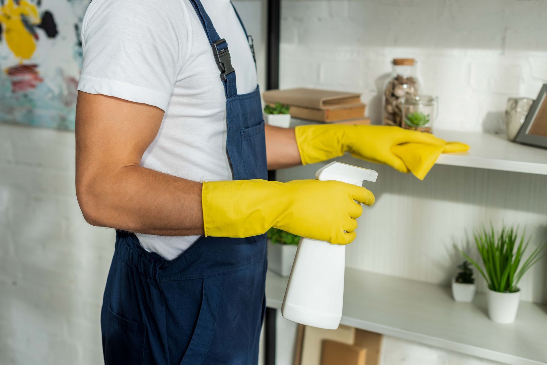 Person wearing yellow gloves, overalls, and white shirt cleaning a shelf with a spray bottle.