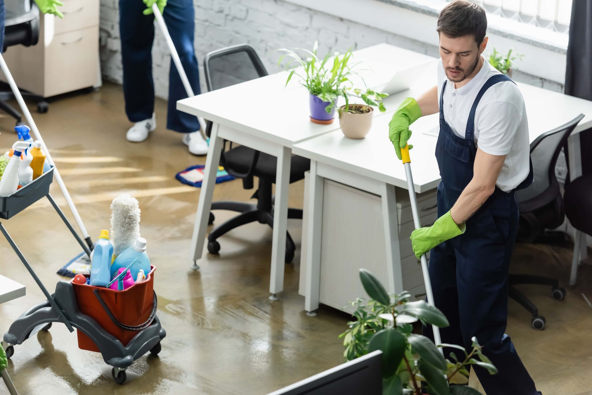 Office cleaning crew mopping a floor; man in overalls and green gloves. A cart with cleaning supplies is nearby.