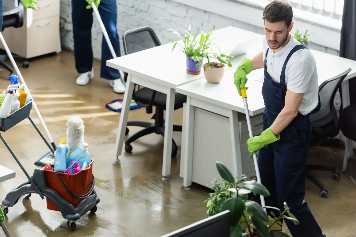 Office cleaners mopping the floor. Men in blue overalls, green gloves, and a cleaning cart.