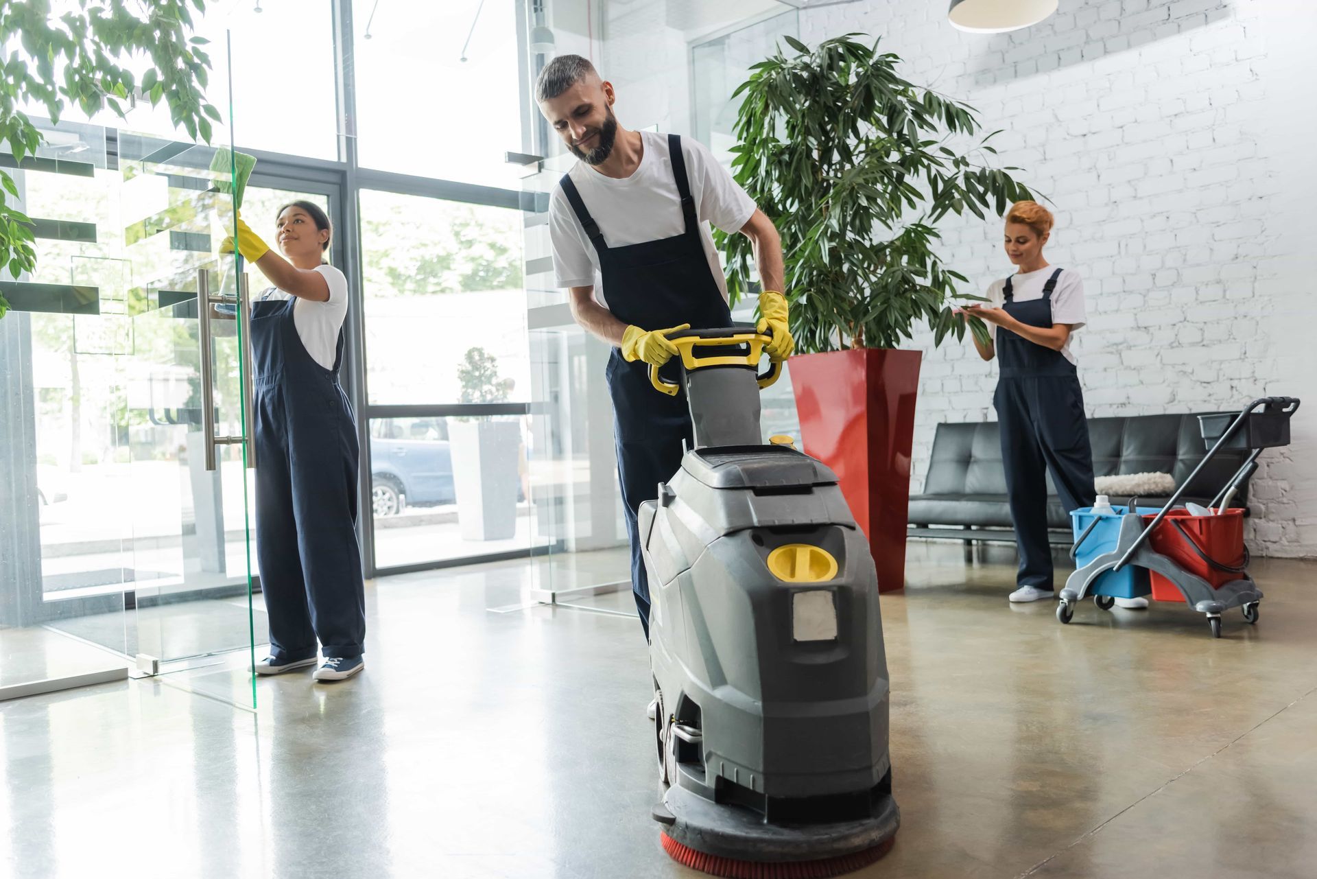 Three cleaners in blue overalls cleaning an office. One operates a floor cleaner, others clean windows and surfaces.