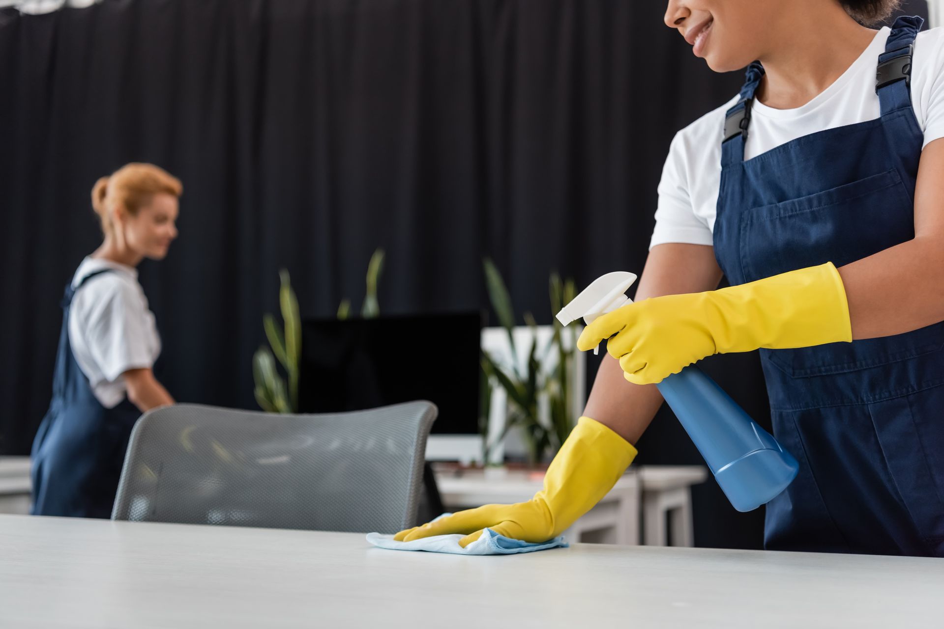 Two women cleaning an office: One sprays, the other tidies in front of a dark backdrop.