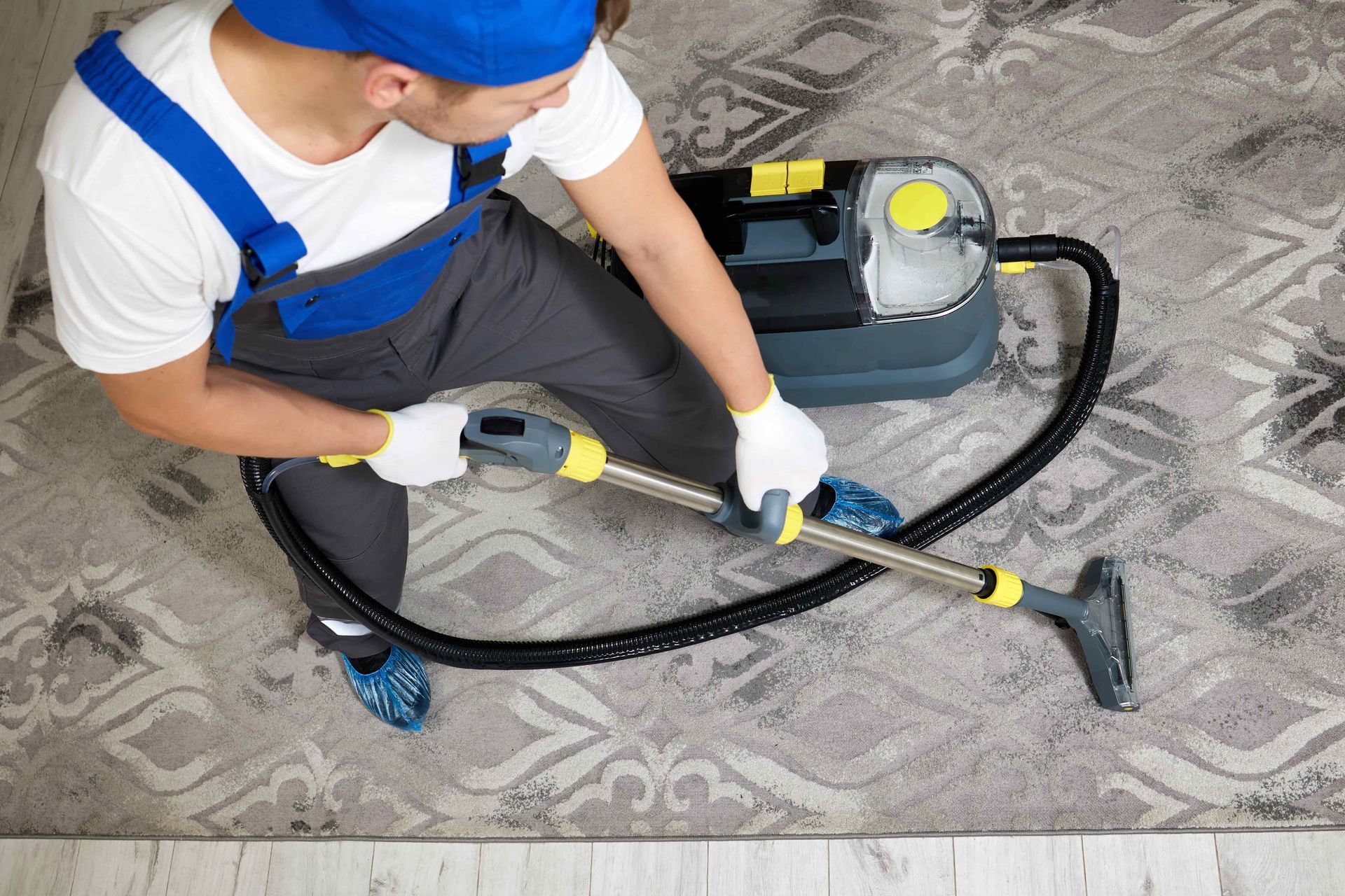 Man cleaning a patterned rug with a carpet cleaner machine, wearing blue overalls and gloves.