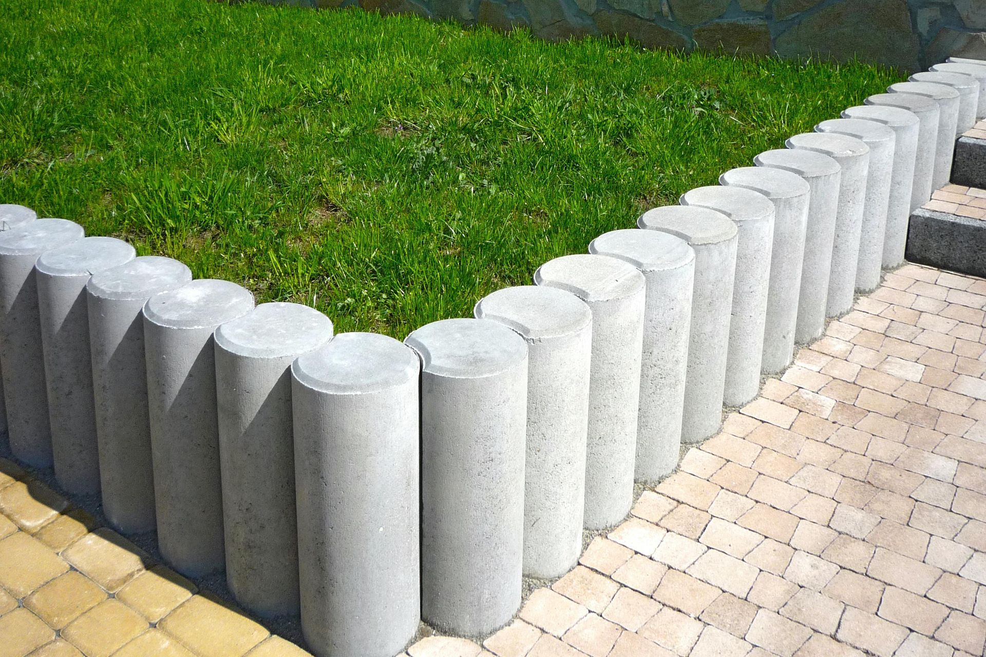 Gray cylindrical concrete border edging a grassy patch next to a paved pathway.