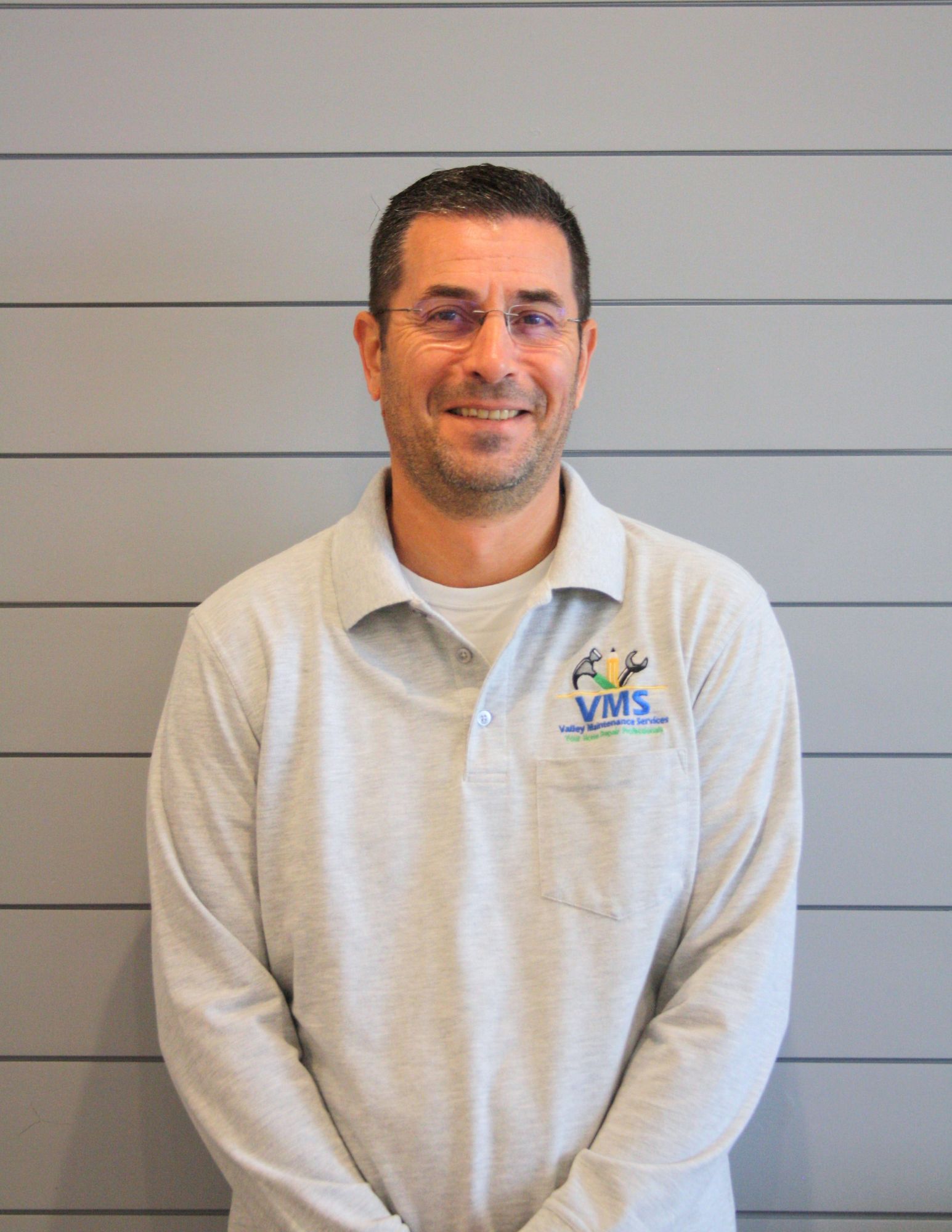 A man wearing a grey shirt with the word vms on it is standing in front of a wall.