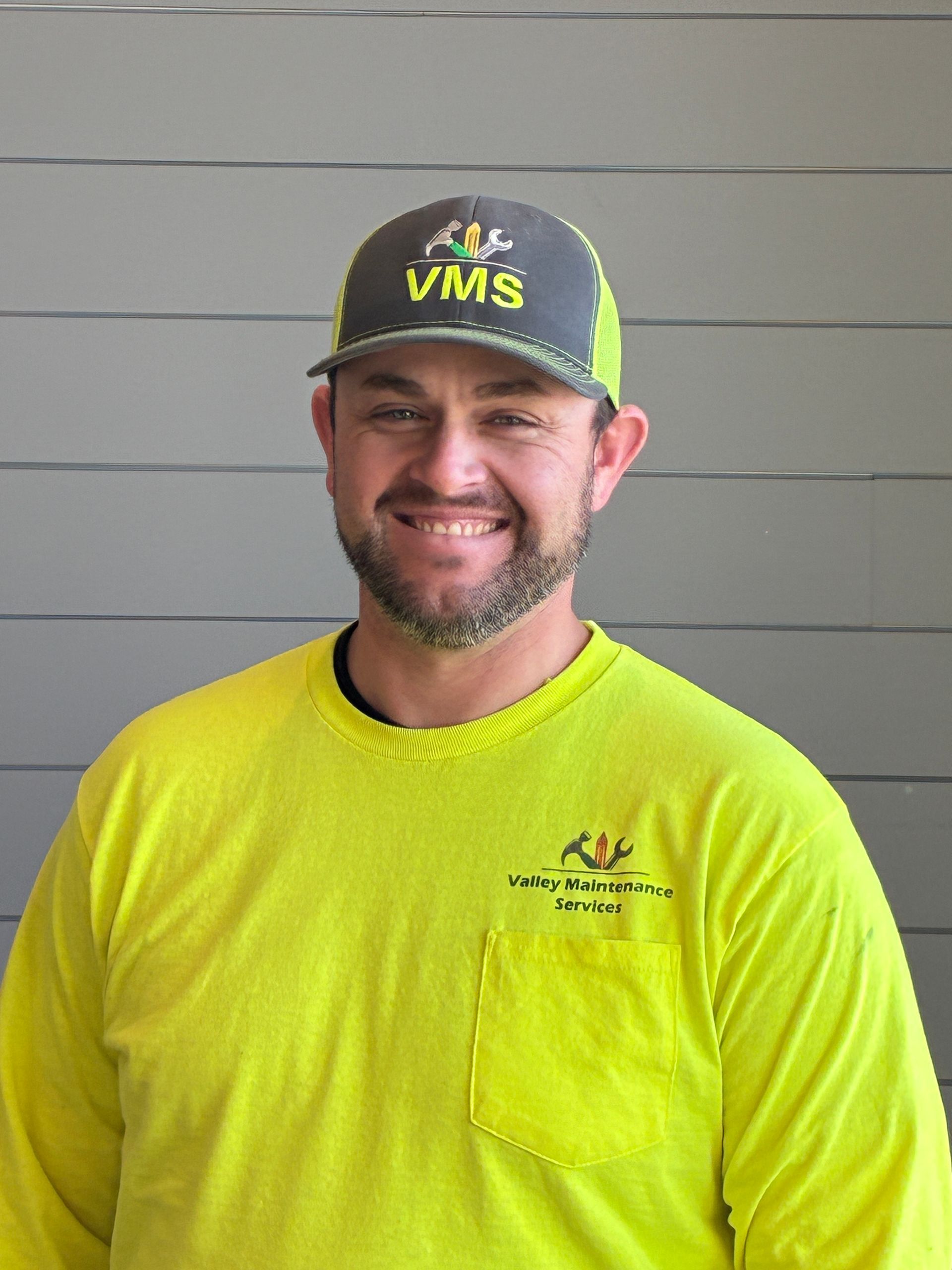 Man in a green work shirt and cap, standing in front of a gray wall.