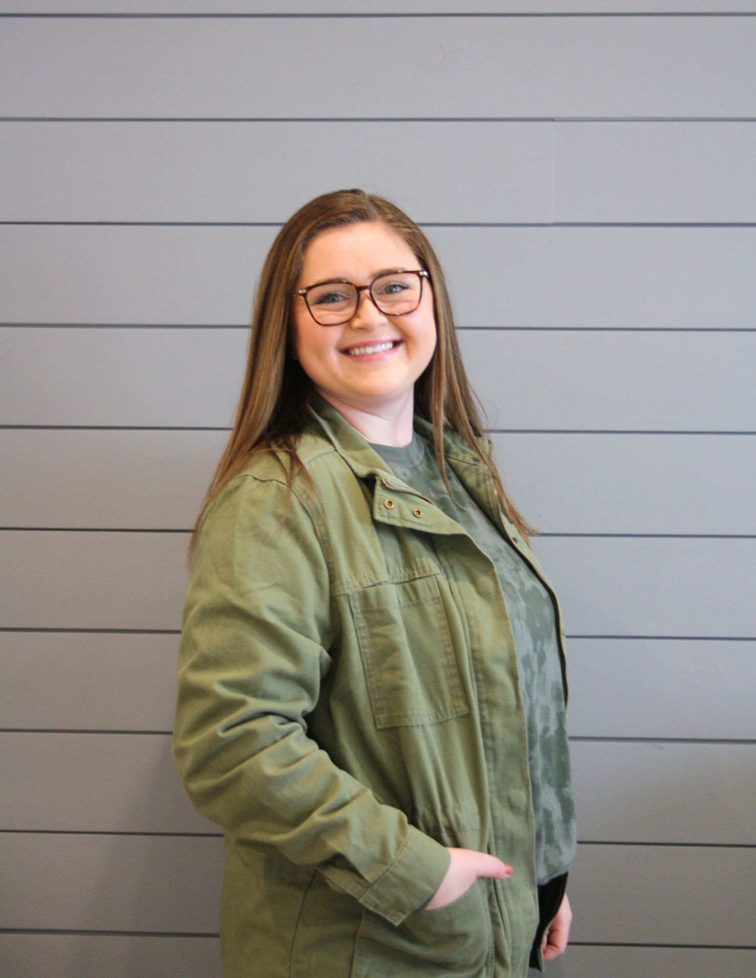 A woman wearing glasses and a green jacket is standing in front of a gray wall.