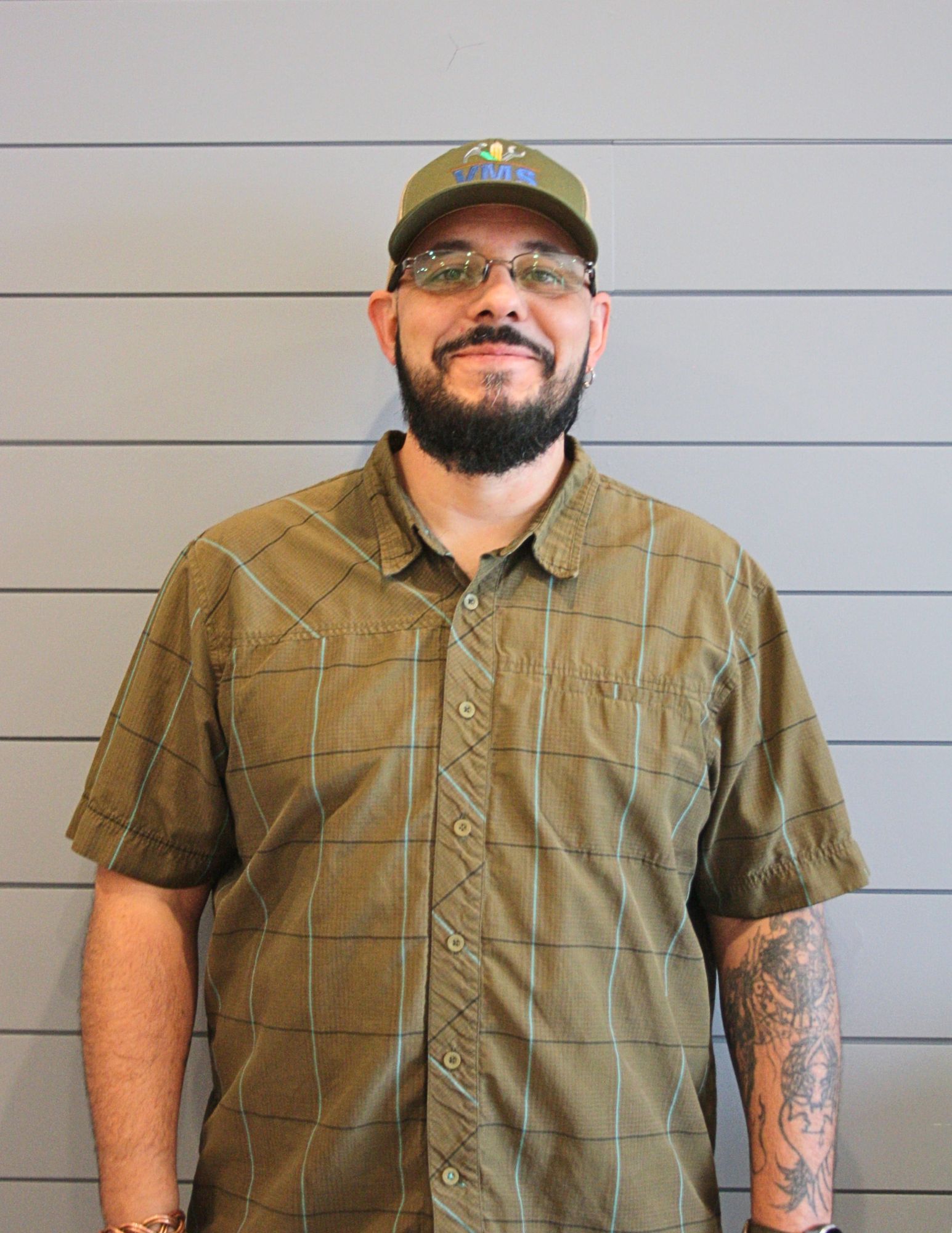 A man with a beard wearing a hat and glasses is standing in front of a wall.