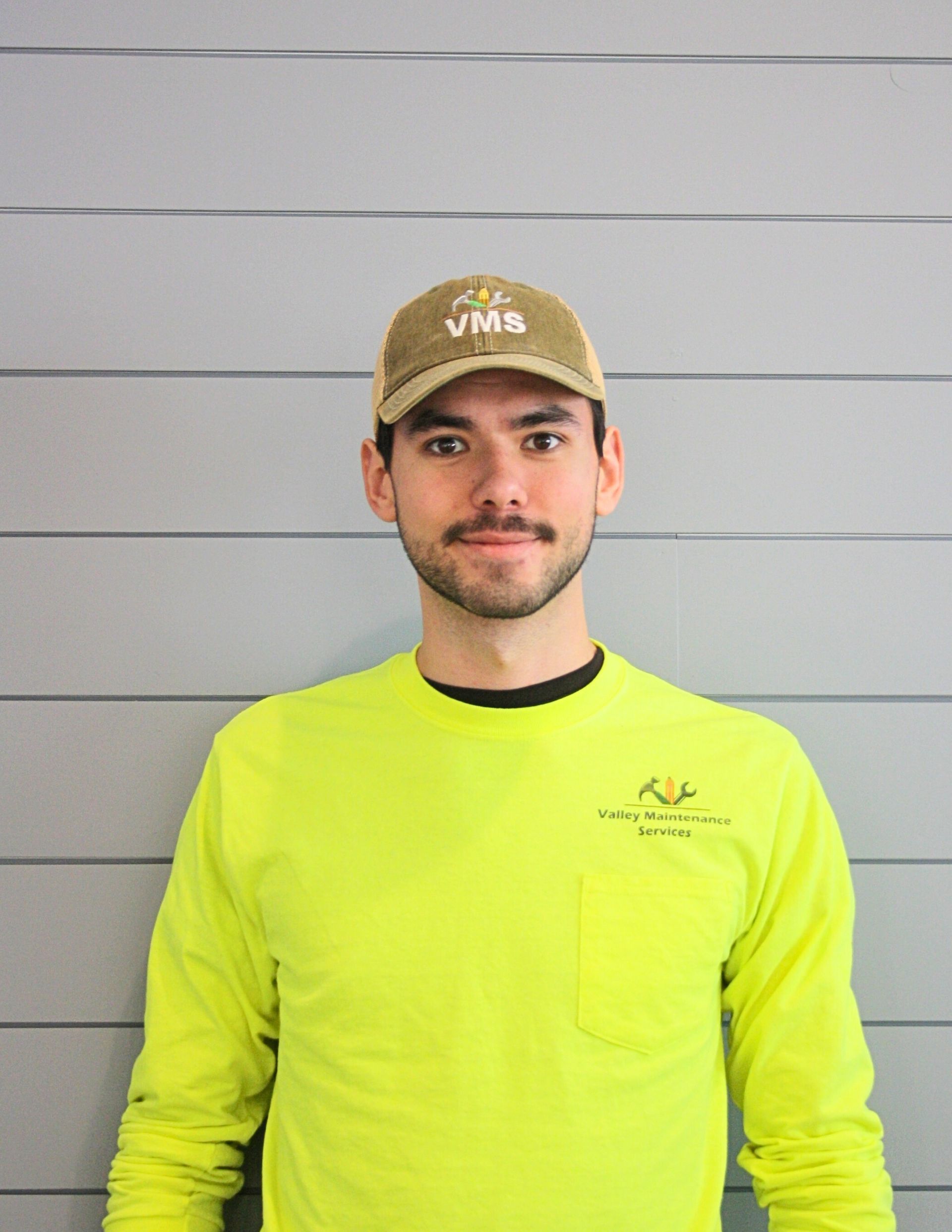 Man in a green work shirt and cap, standing in front of a gray wall.