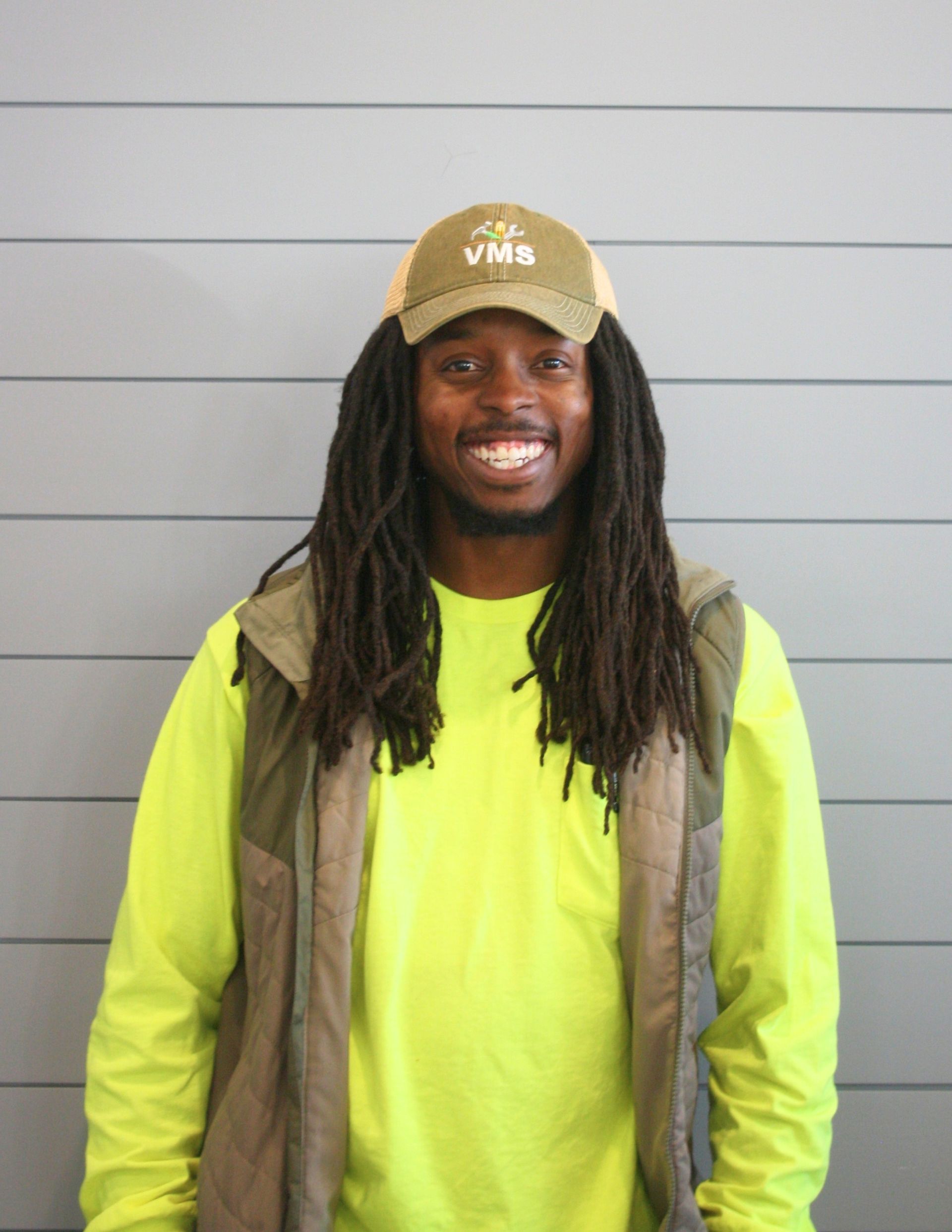 A smiling man with dreadlocks wears a tan cap, safety vest, and yellow long-sleeve shirt, standing in front of a gray wall.