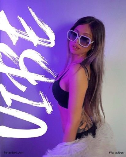 Woman wearing sunglasses and a black top, posing by a purple wall with white graffiti.