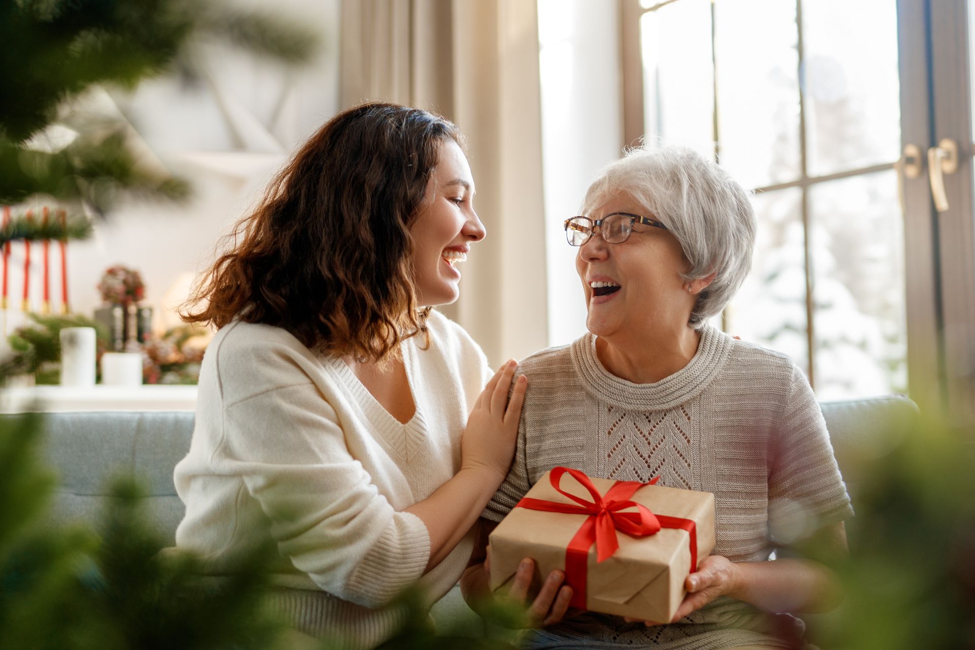 Woman giving a gift to an older woman with a happy expression, seated indoors with a window in the background.