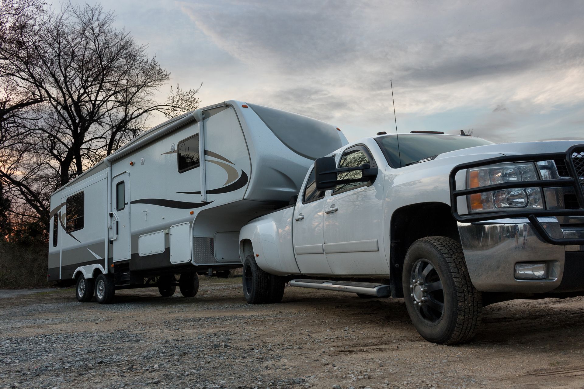 A white truck is towing a trailer in a gravel lot.