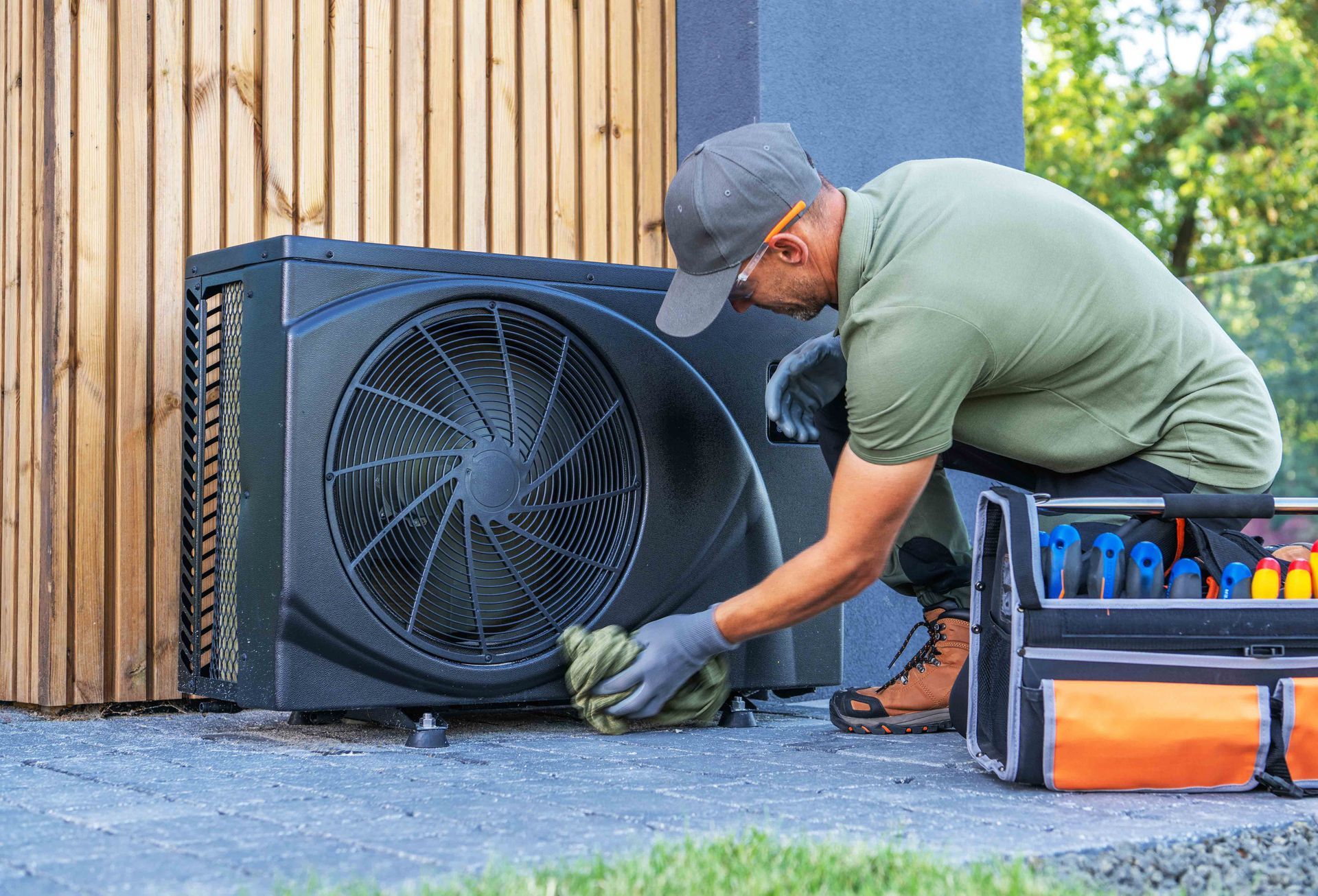 Man inspecting a black heat pump, outdoors. A toolbox is nearby.
