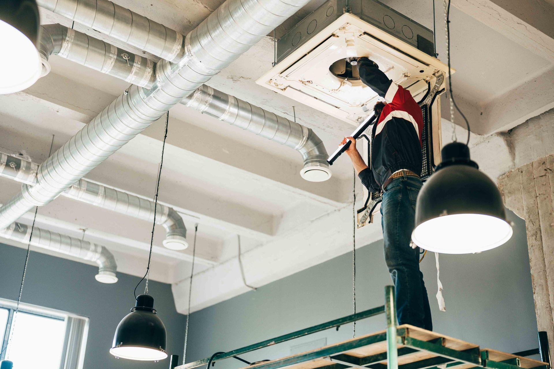 Person on a scaffold repairing ceiling air conditioning unit in a room with industrial design elements.