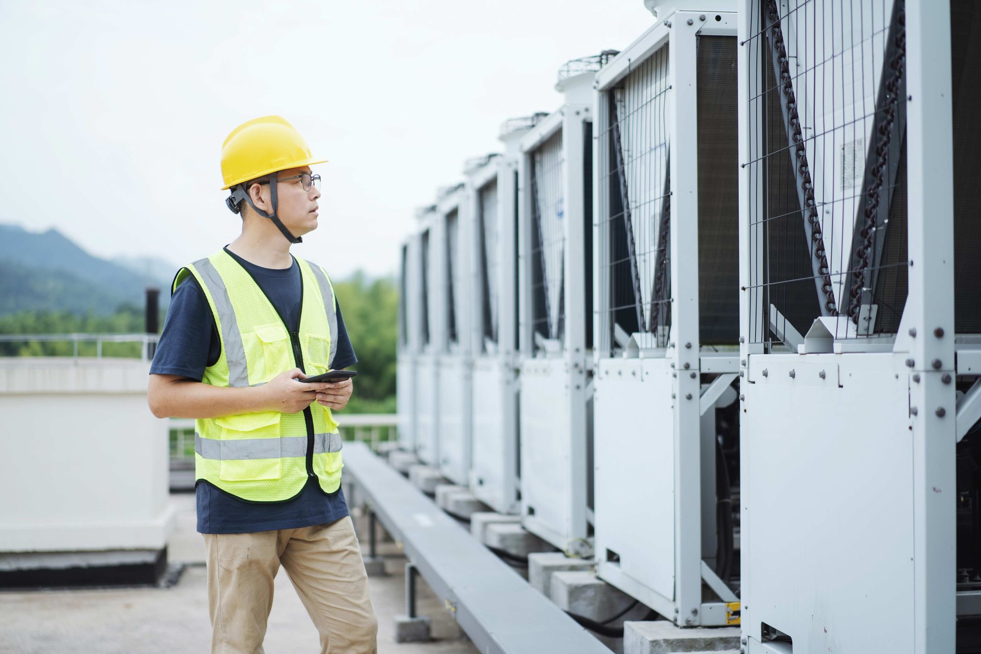 Technician in yellow vest and hard hat inspects HVAC units on a rooftop, holding a tablet.