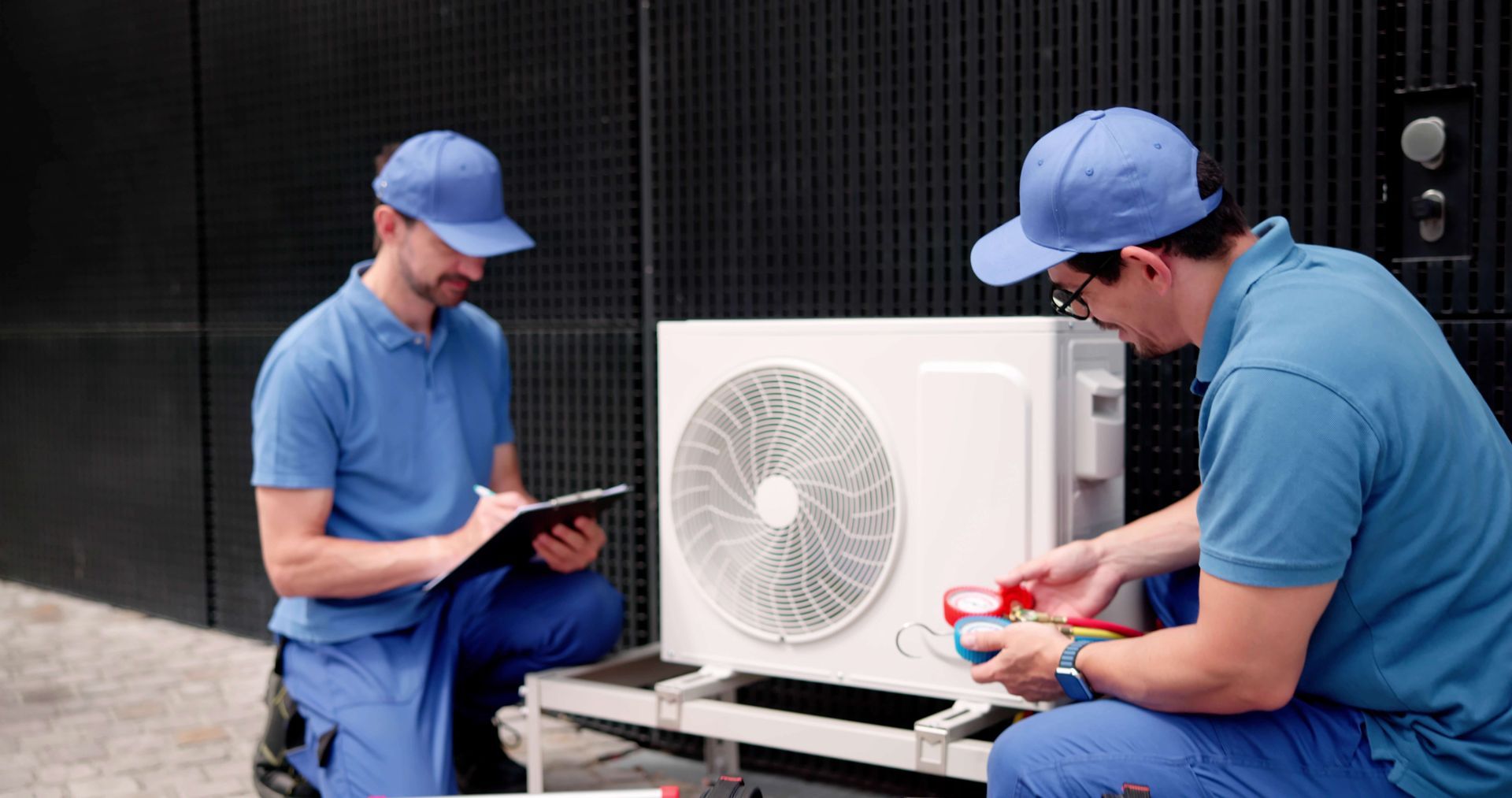Two HVAC technicians in blue uniforms work on an air conditioning unit. One checks a clipboard, the other attaches gauges.