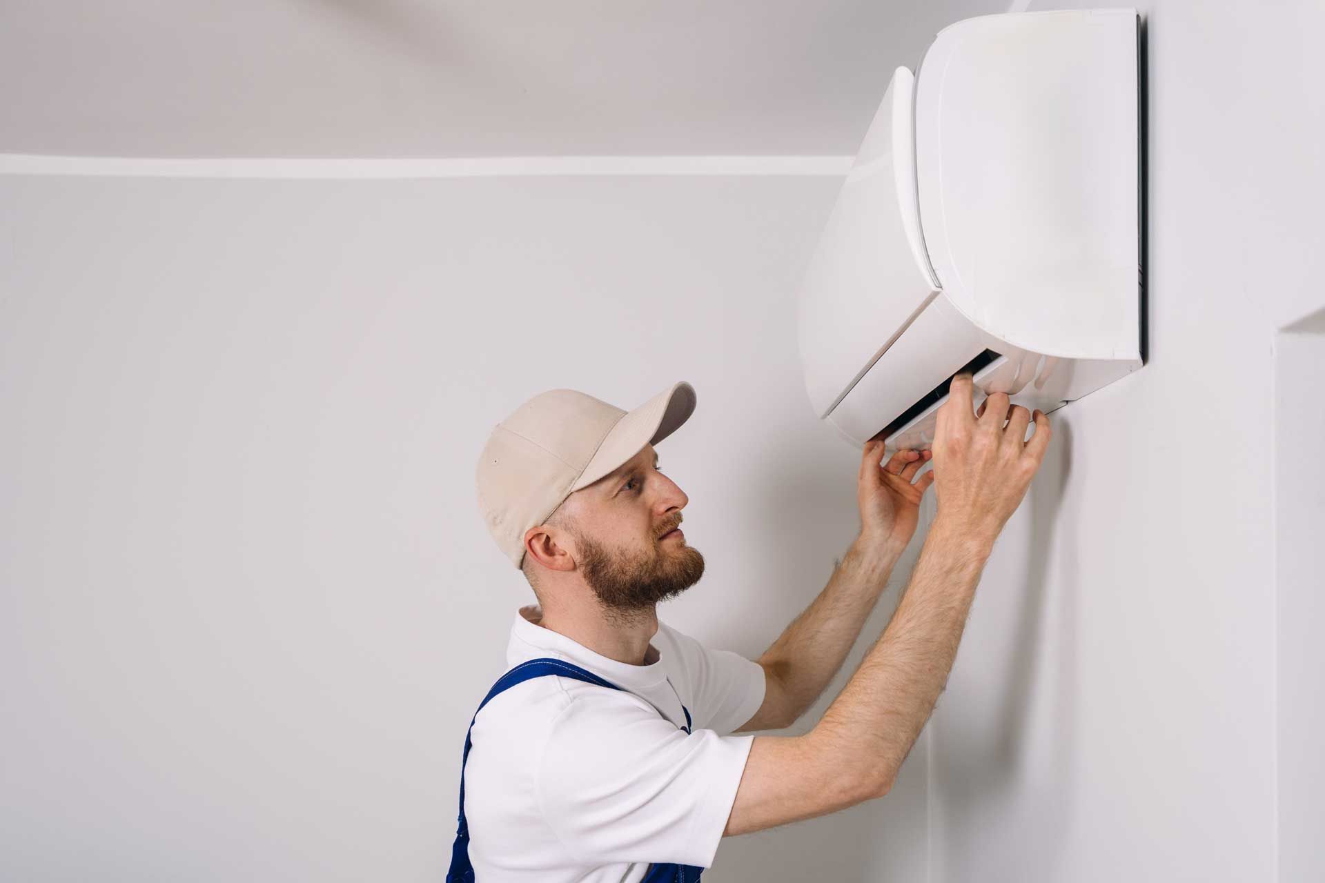 Technician in cap and overalls fixing a wall-mounted air conditioning unit in a room with white walls.