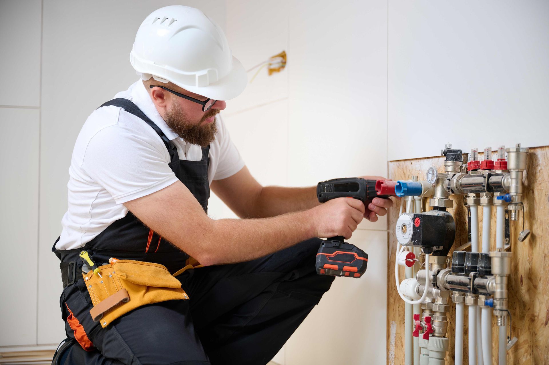 Plumber in overalls, hard hat, using a drill on pipes and valves attached to a wall.