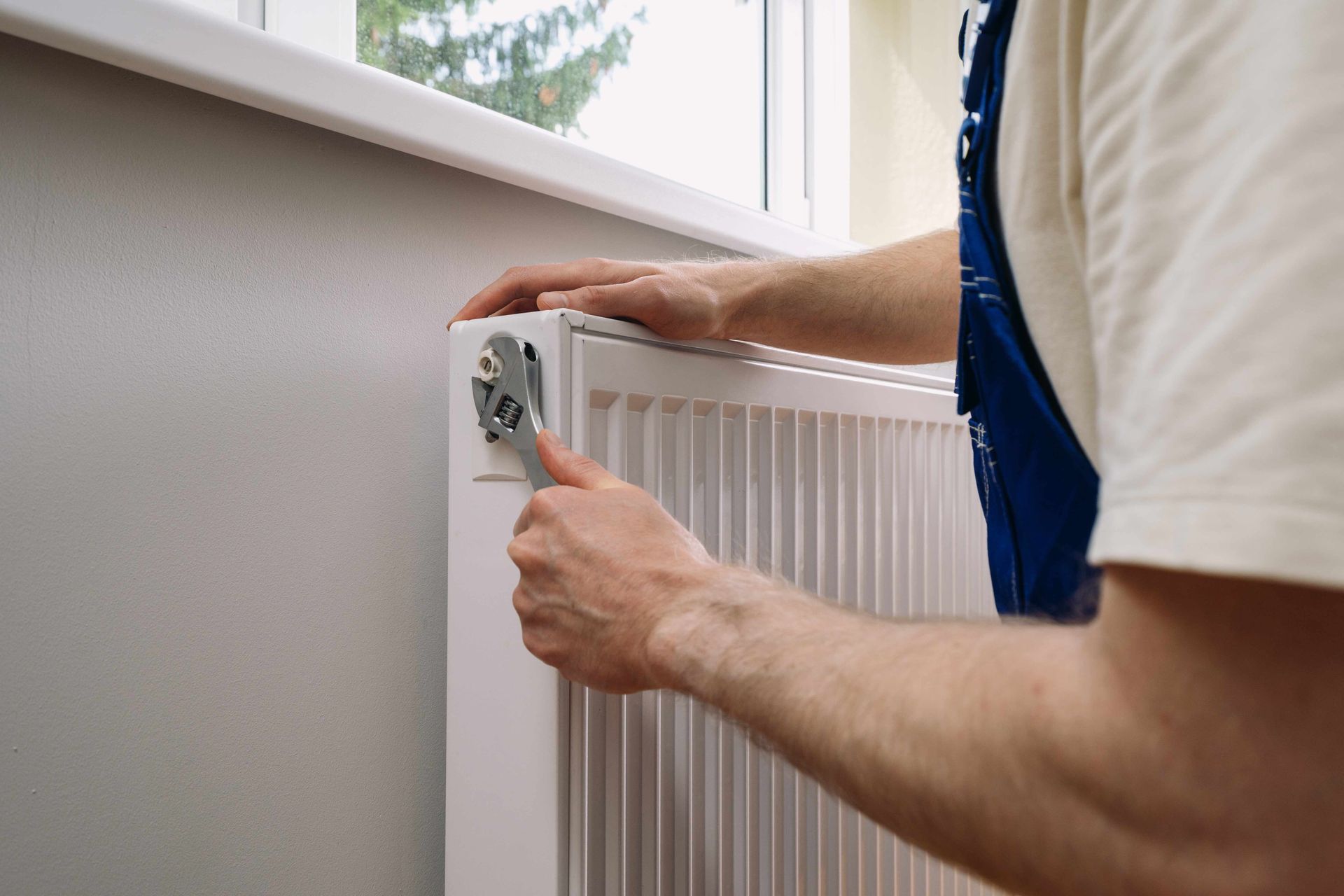 Person using a wrench to adjust a white radiator near a window.