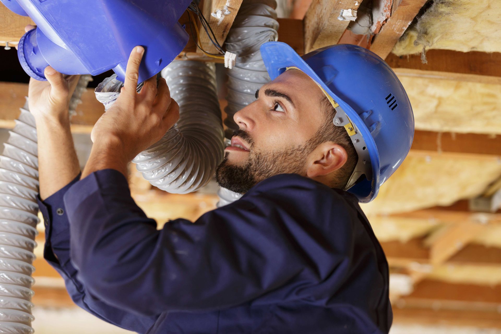 Man in blue hard hat works on ductwork in a home attic.