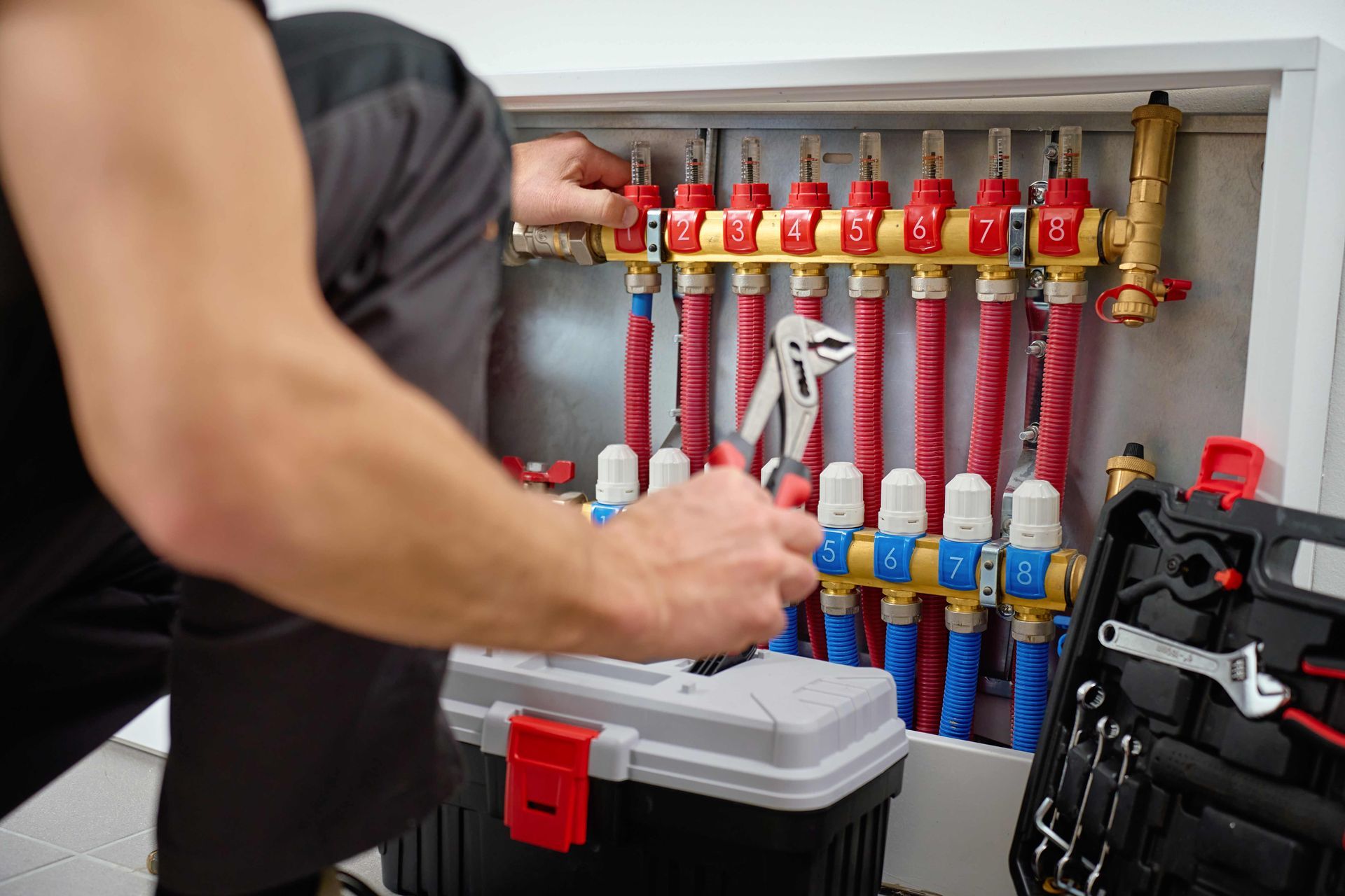 Plumber using pliers to work on a radiant floor heating manifold in a white utility space.