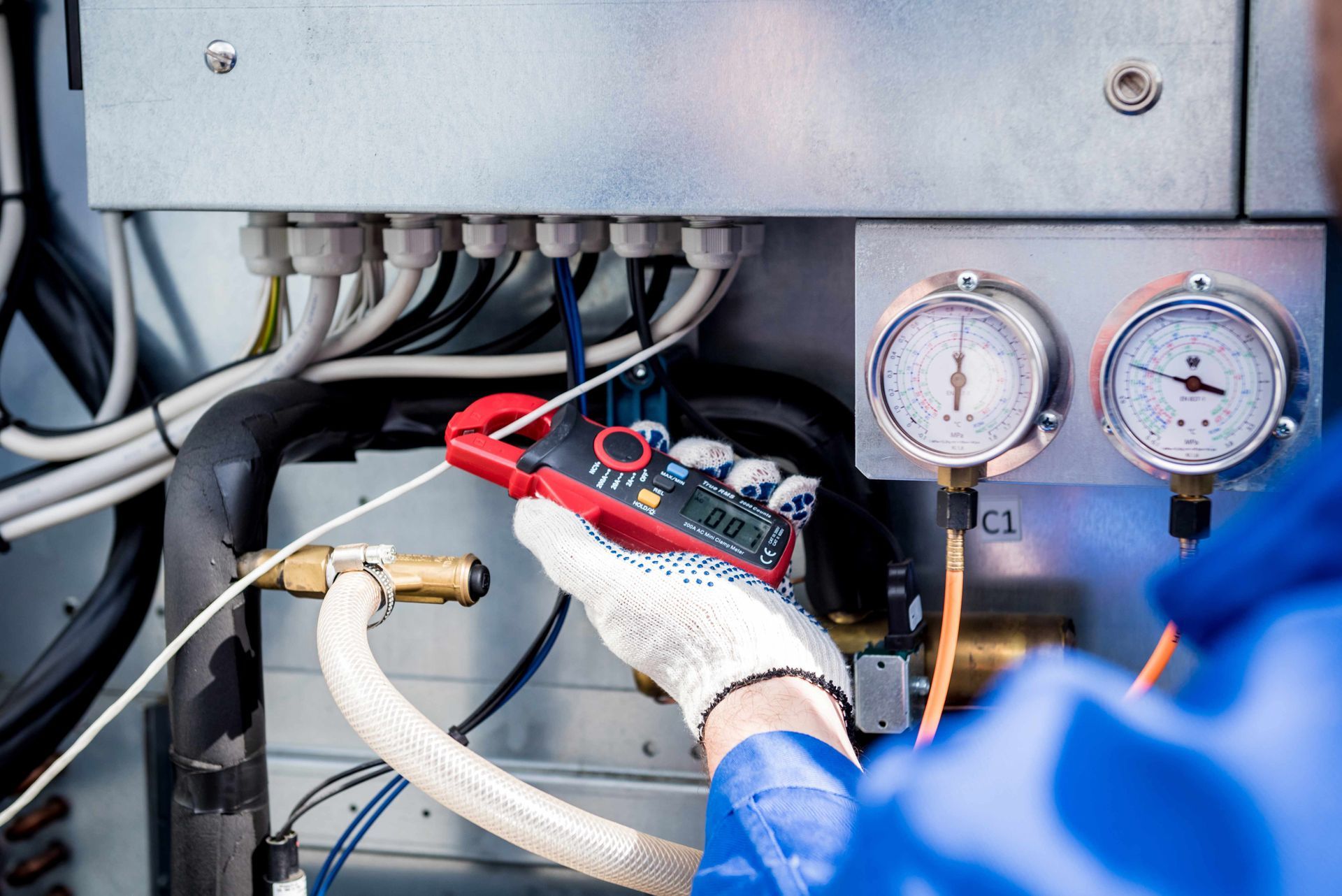 Person in blue uniform using a red clamp meter on wires inside a metal electrical panel. Gauges and pipes are also visible.