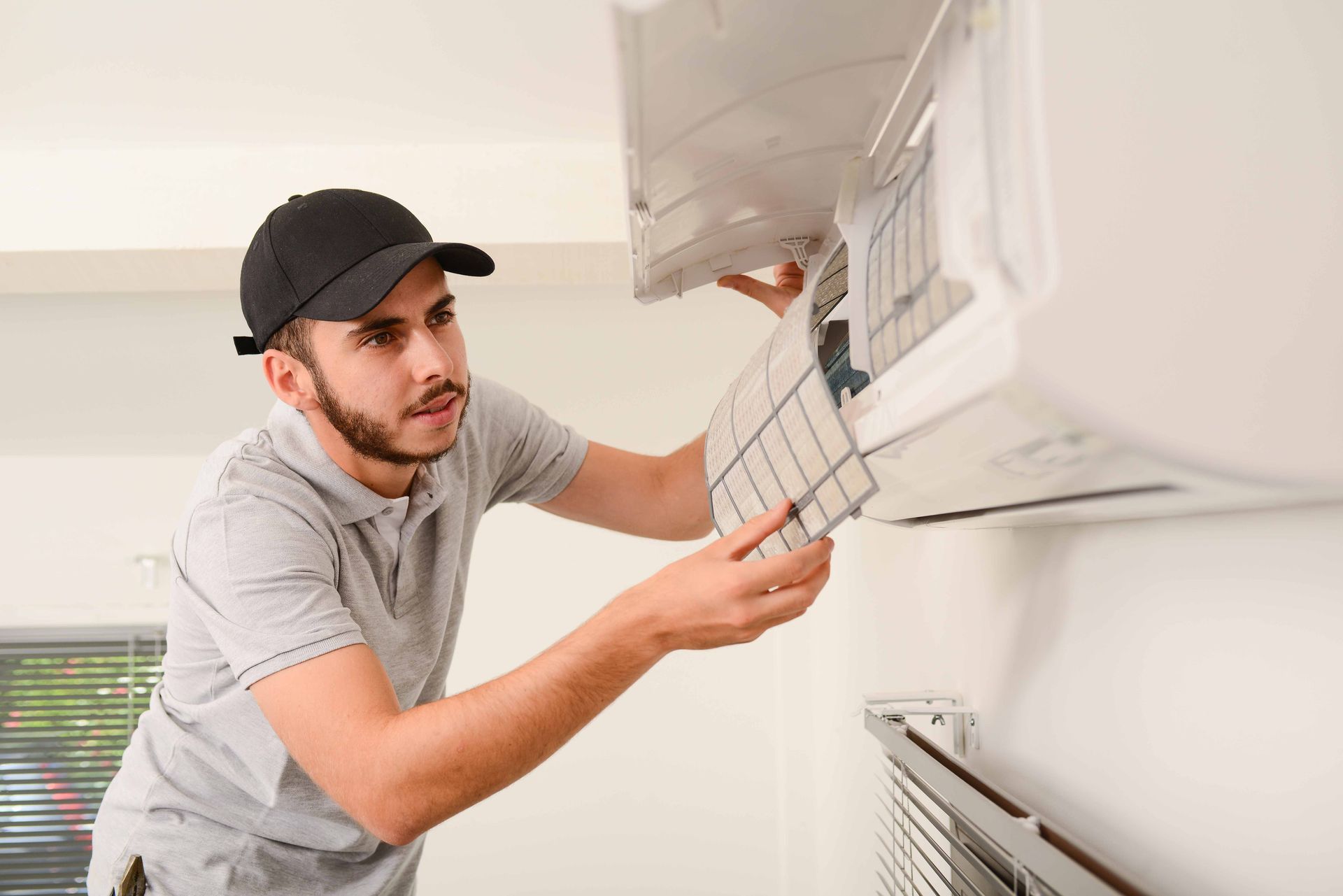 HVAC technician in black cap, removing AC filter from wall unit, indoors.