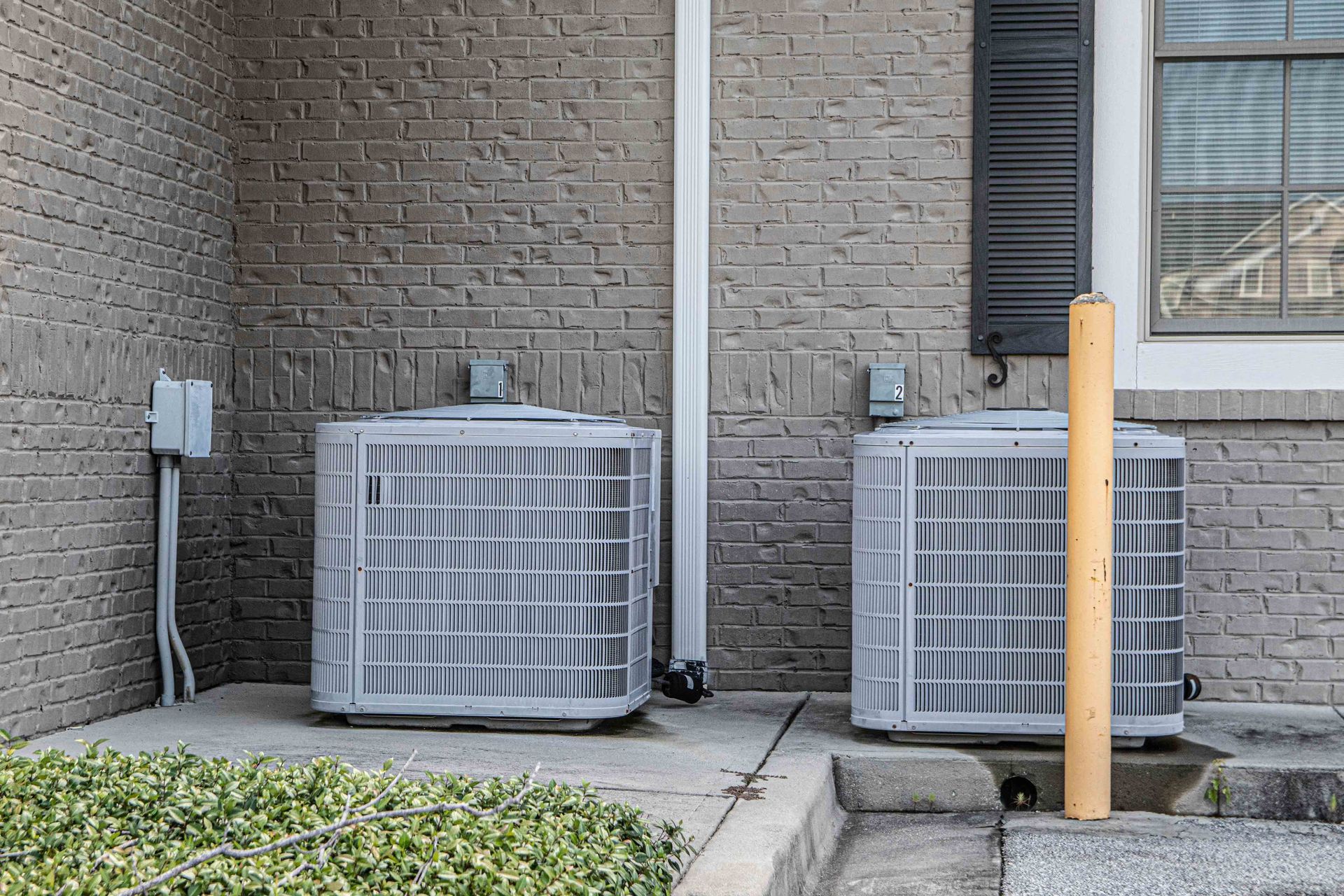 Two grey air conditioning units next to a brick building.