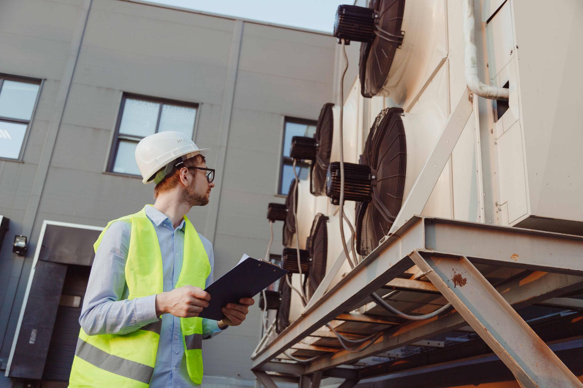 HVAC technician in hardhat and vest inspecting rooftop air conditioning units, holding clipboard.