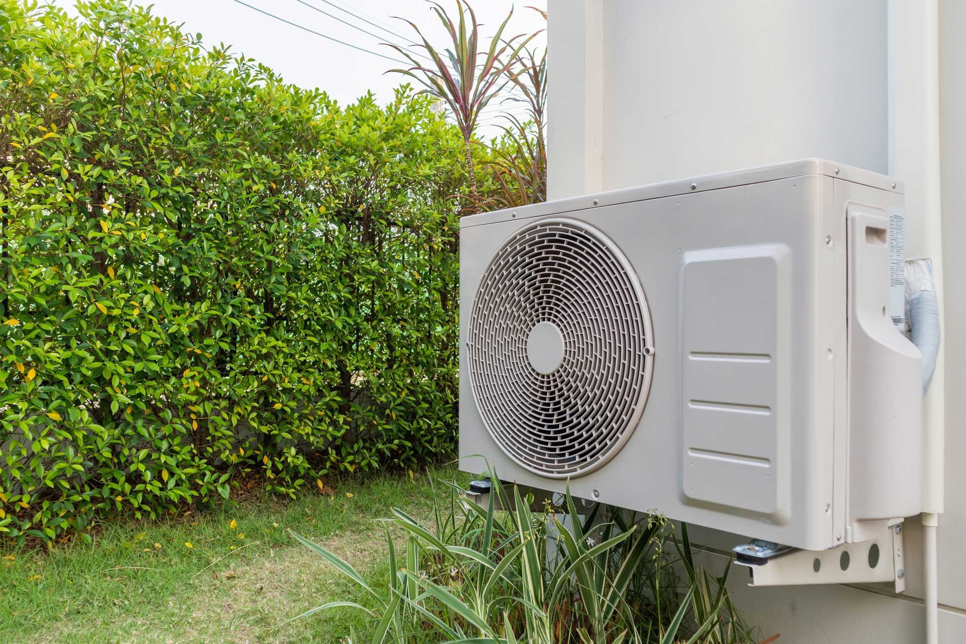 Air conditioning unit mounted on a white building wall next to green foliage.