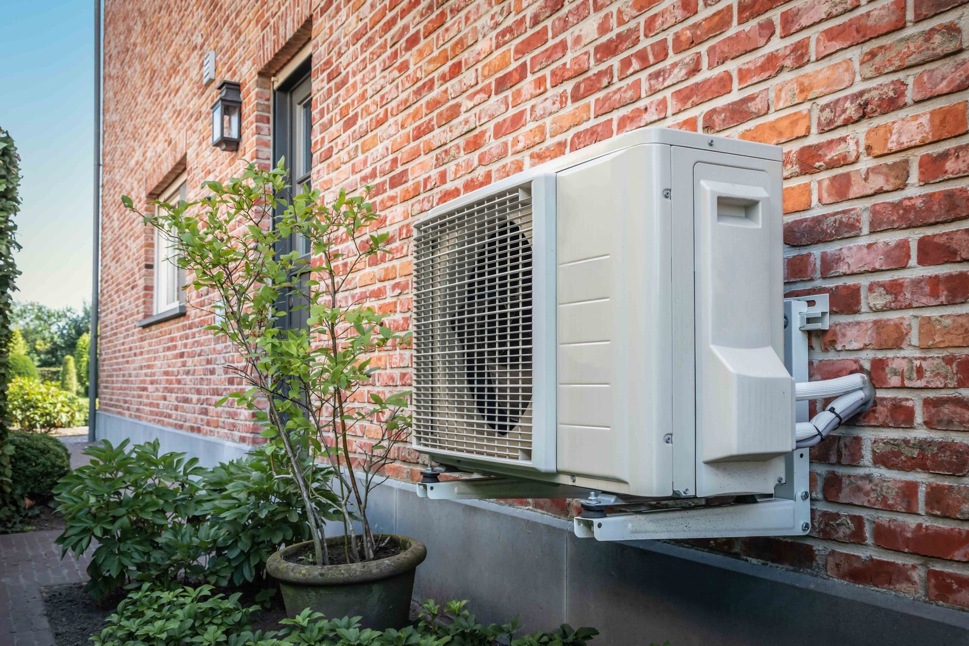 Heat pump mounted on a brick building exterior, near a potted plant.