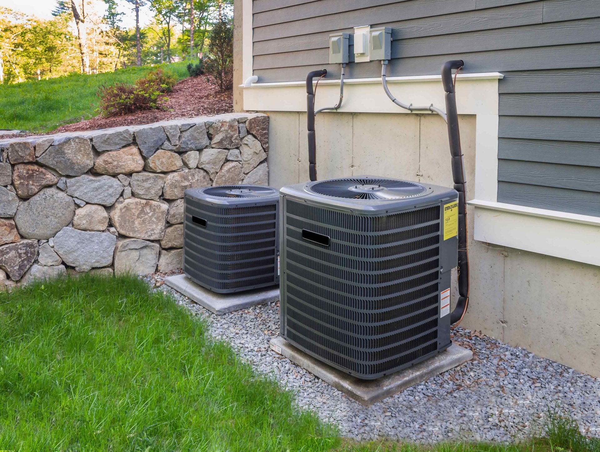 Two outdoor air conditioning units on concrete pads near a stone wall and house siding.