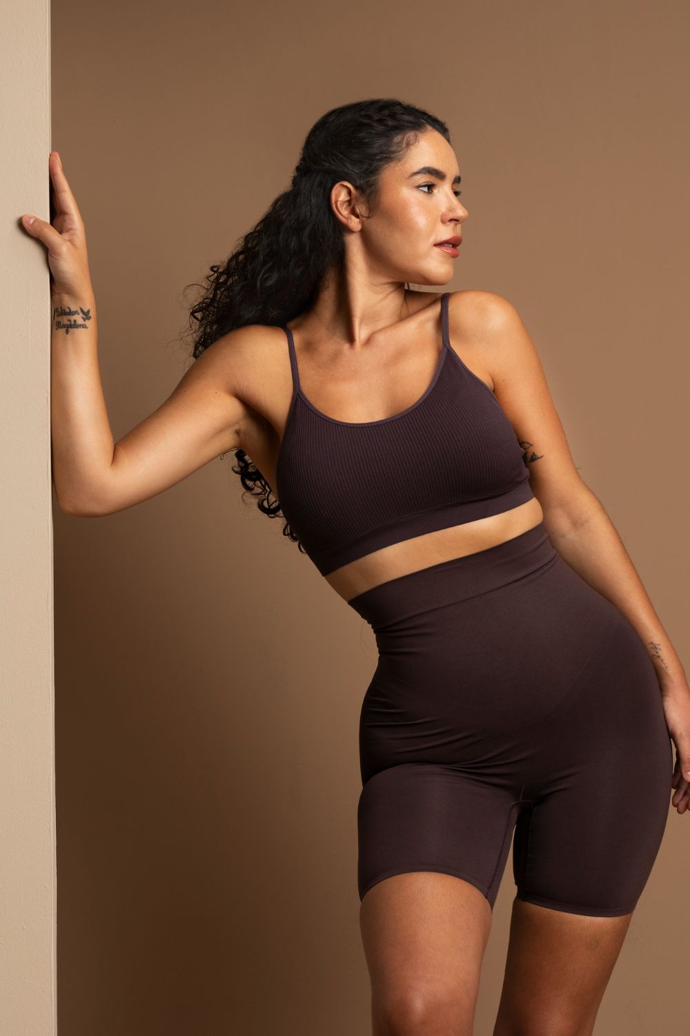 Mujer con un conjunto deportivo de color marrón, apoyada contra una pared, mirando hacia un lado.