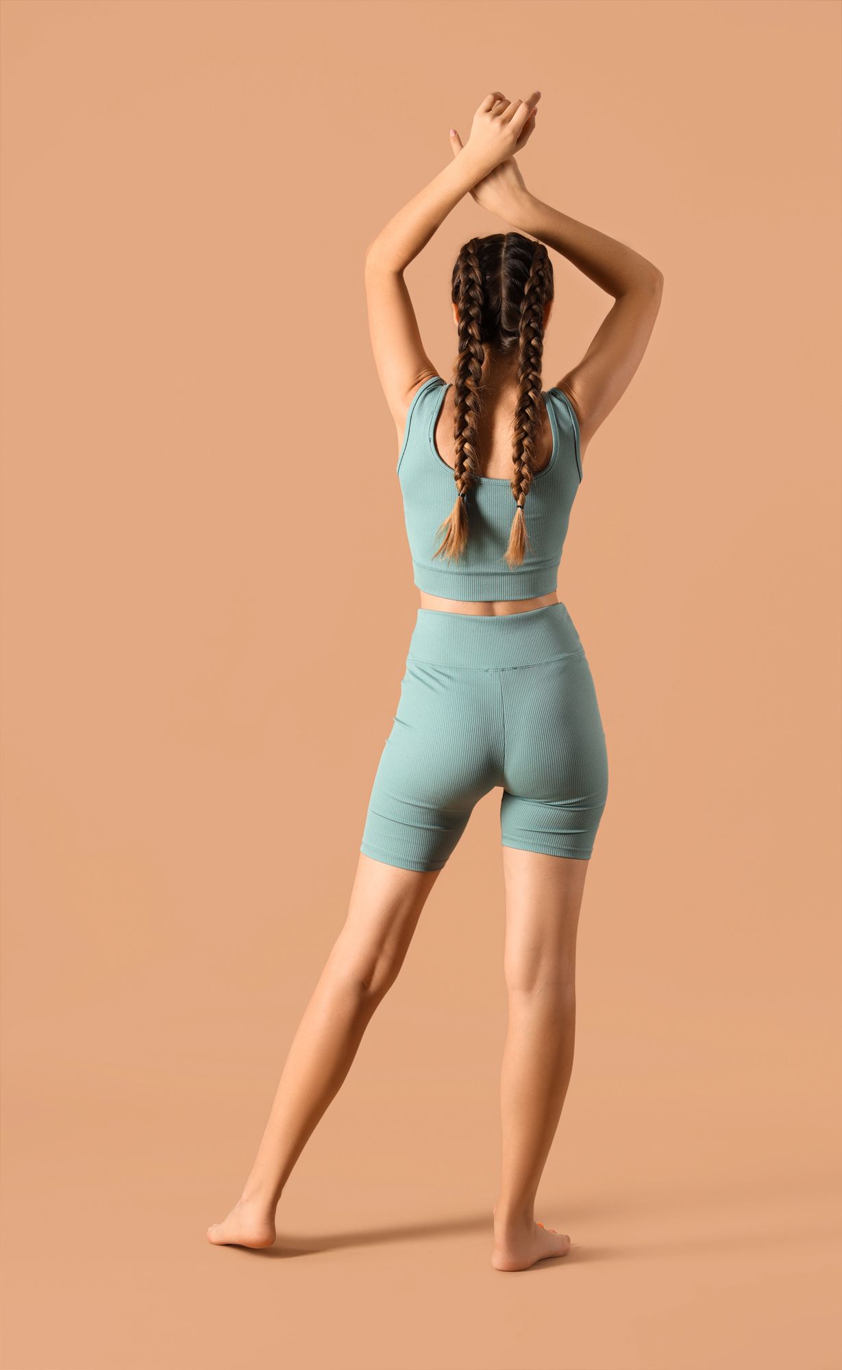 Mujer con ropa deportiva color verde azulado, brazos levantados hacia arriba, trenzas, fondo neutro.