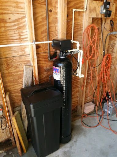 A water softener is sitting in a garage next to a trash can.