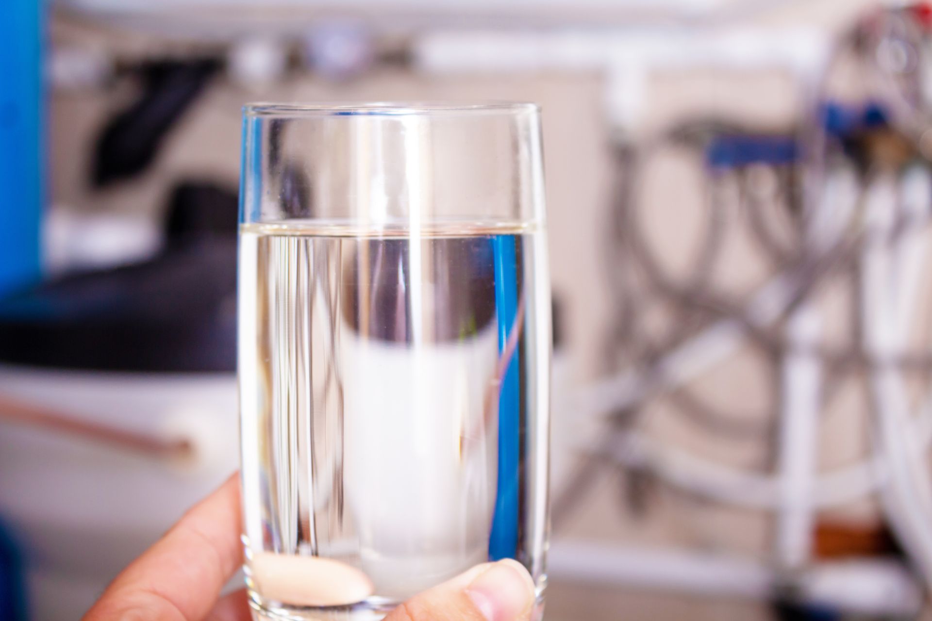 Hand holding a clear glass of filtered water with home water treatment equipment blurred behind.