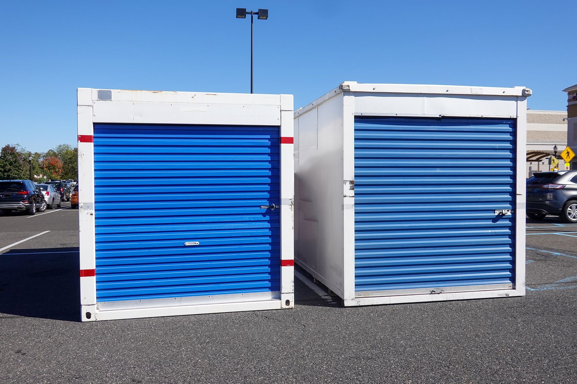 Two white storage containers with blue roll-up doors in a parking lot on a sunny day.