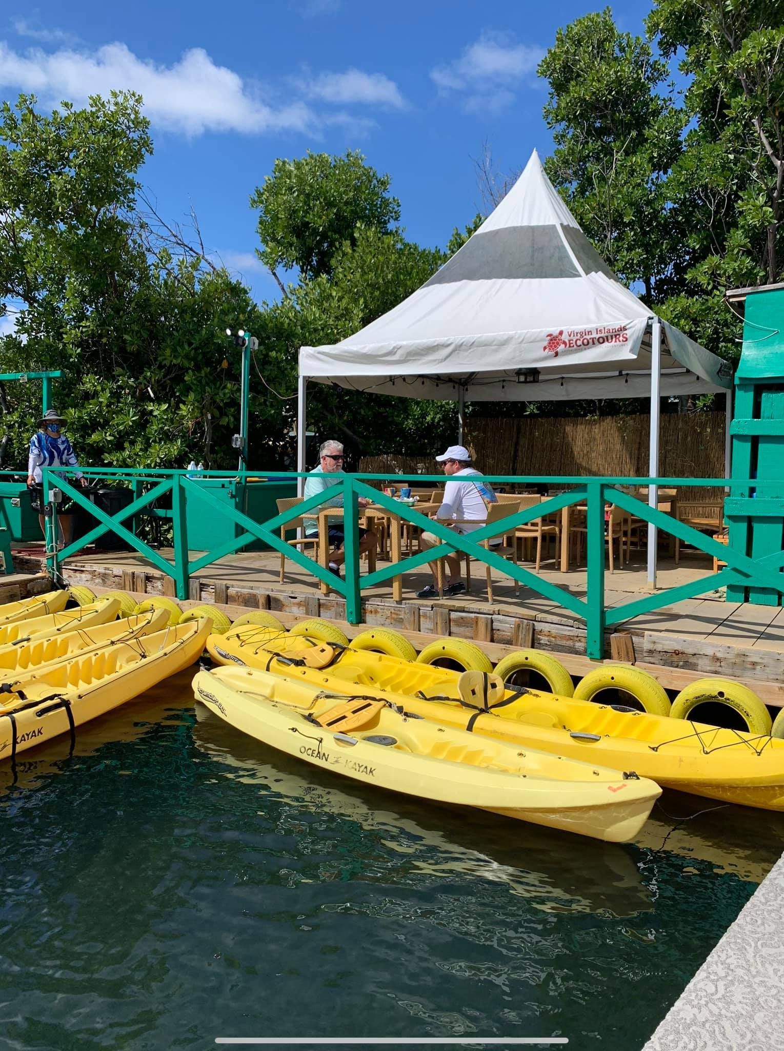 A row of yellow kayaks are sitting in the water near a green fence