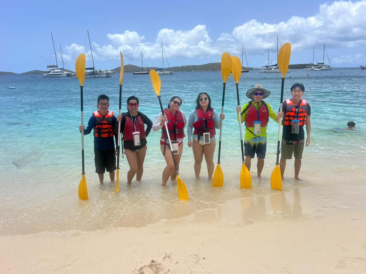 Group of people on a beach holding yellow paddles, wearing life vests, with boats in the background.