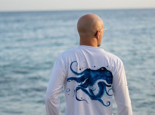 A man wearing a white shirt with an octopus on the back is standing in front of the ocean.