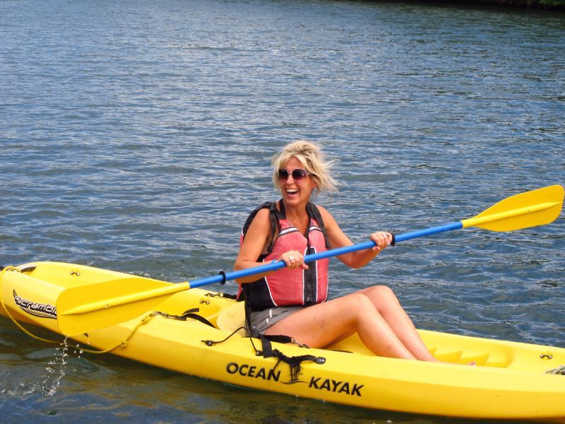 Woman kayaking on calm water; yellow kayak, blue paddle, wearing life vest, sunny day.