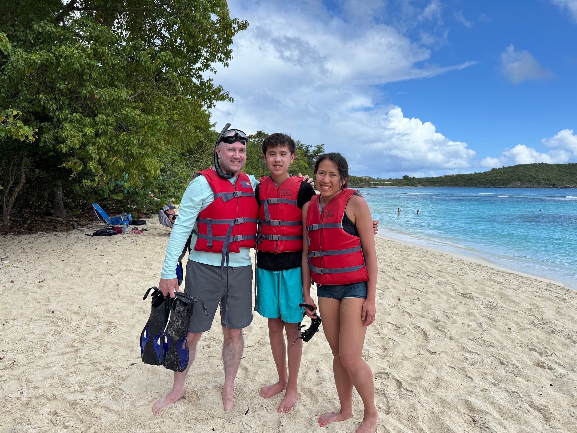 Family in red life vests on a beach, blue water, sunny sky.