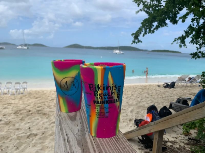 Two colorful cups are sitting on a wooden railing in front of a beach.