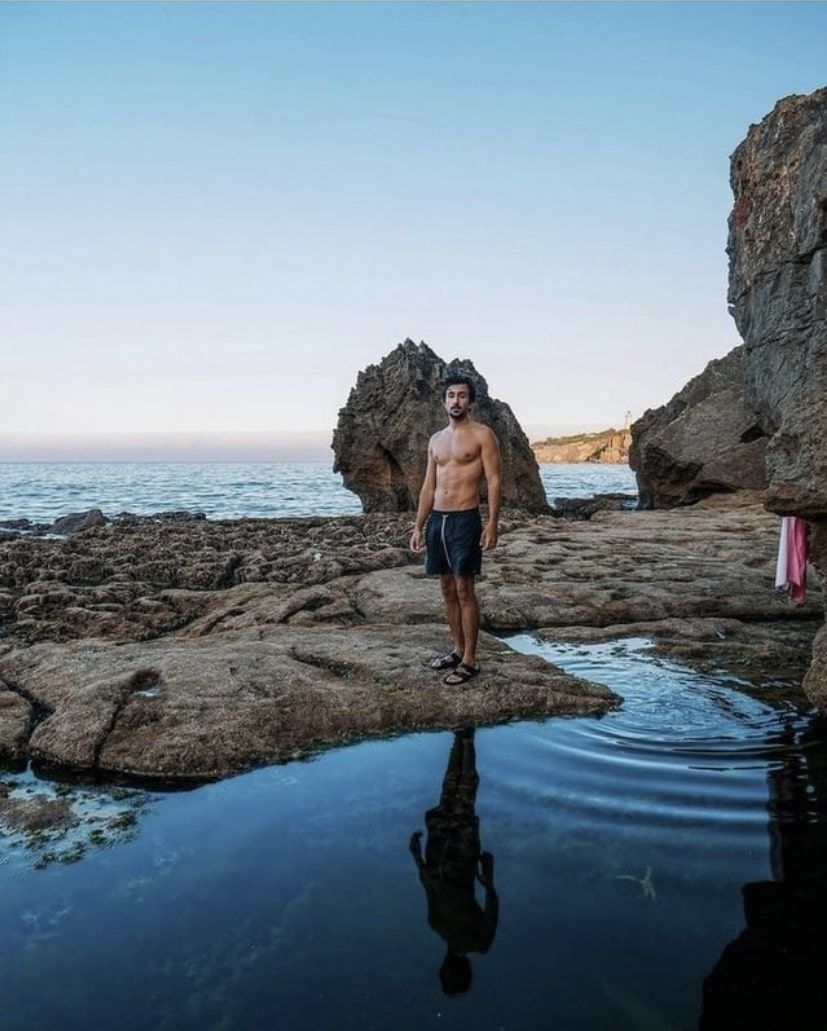 A shirtless man is standing on a rocky beach next to a body of water.