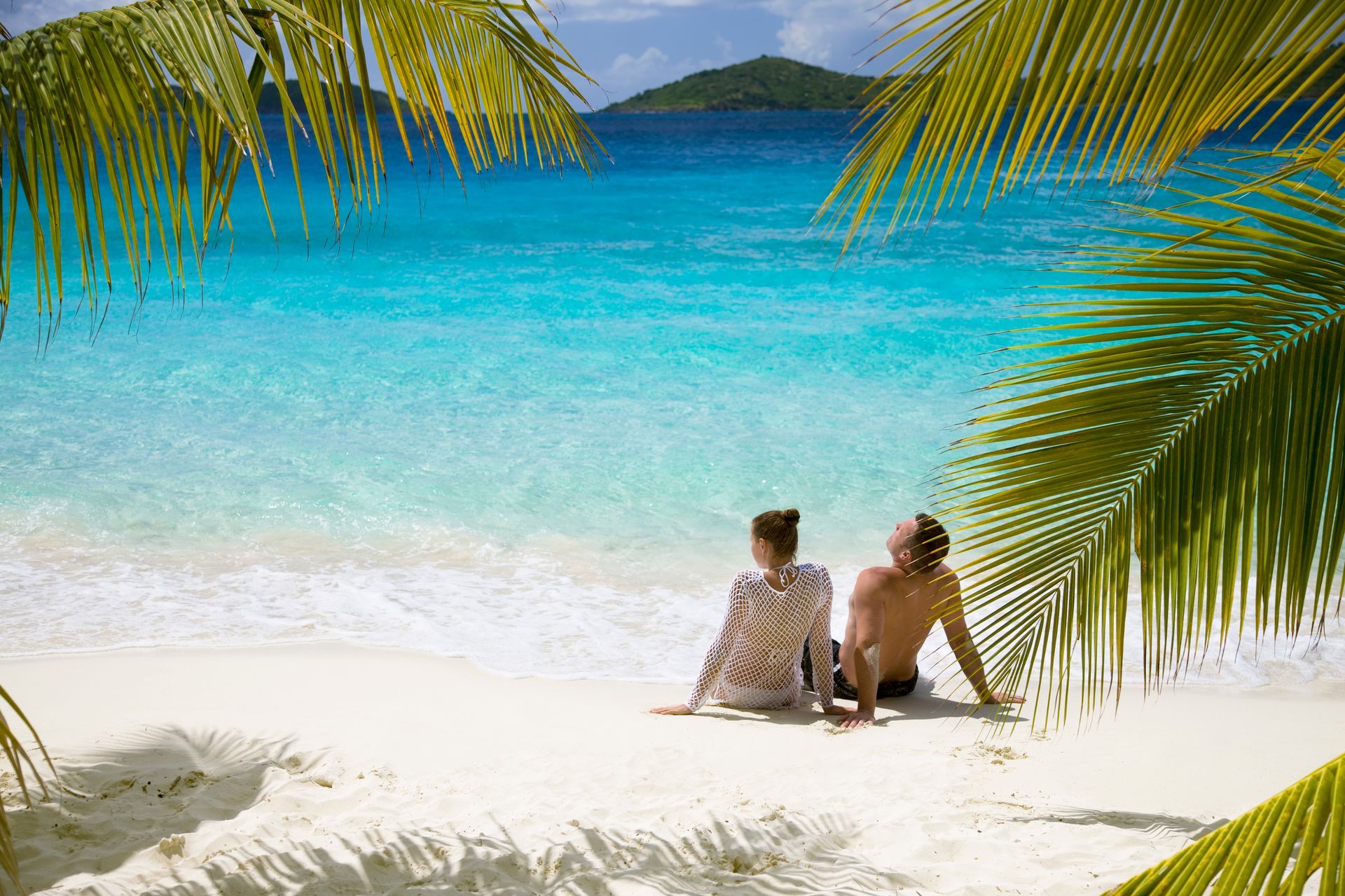 Couple relaxing on white sand beach, turquoise water, palm trees.