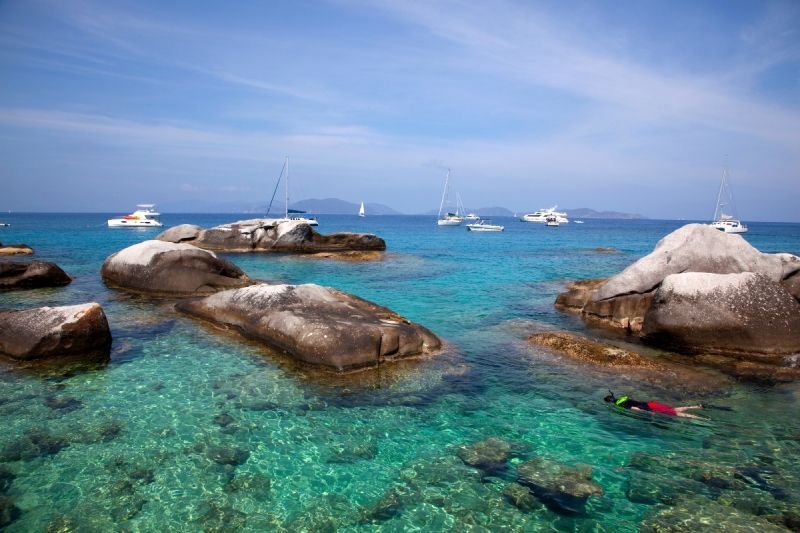 A person is swimming in the ocean surrounded by rocks and boats.