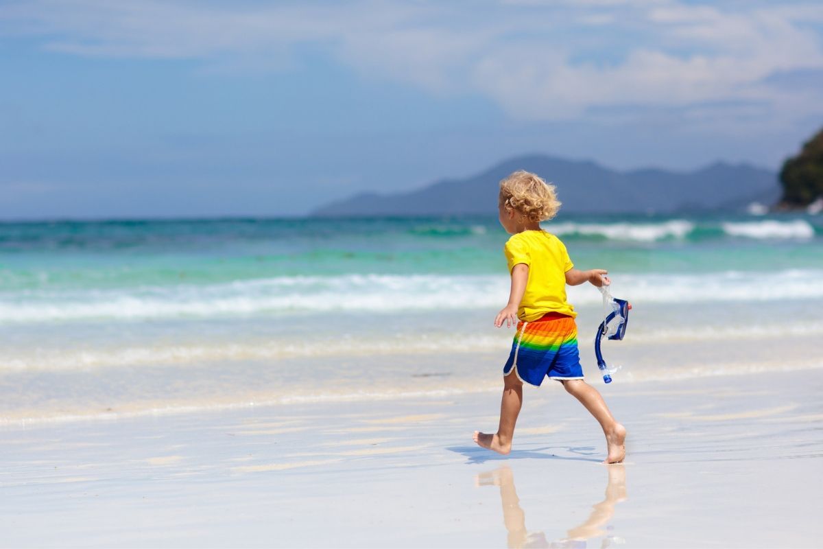 A young boy is walking on the beach holding a pair of goggles.