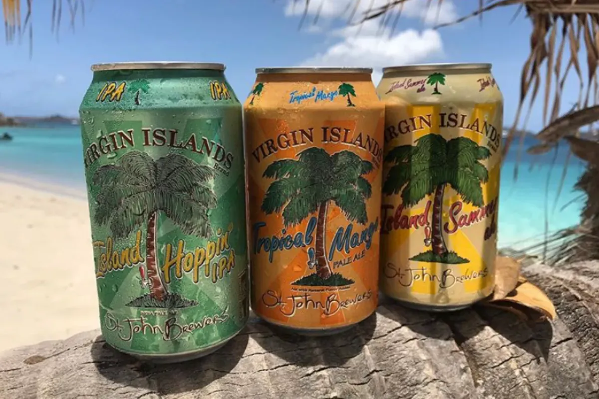 Three cans of virgin island beer are sitting on a log on a beach.