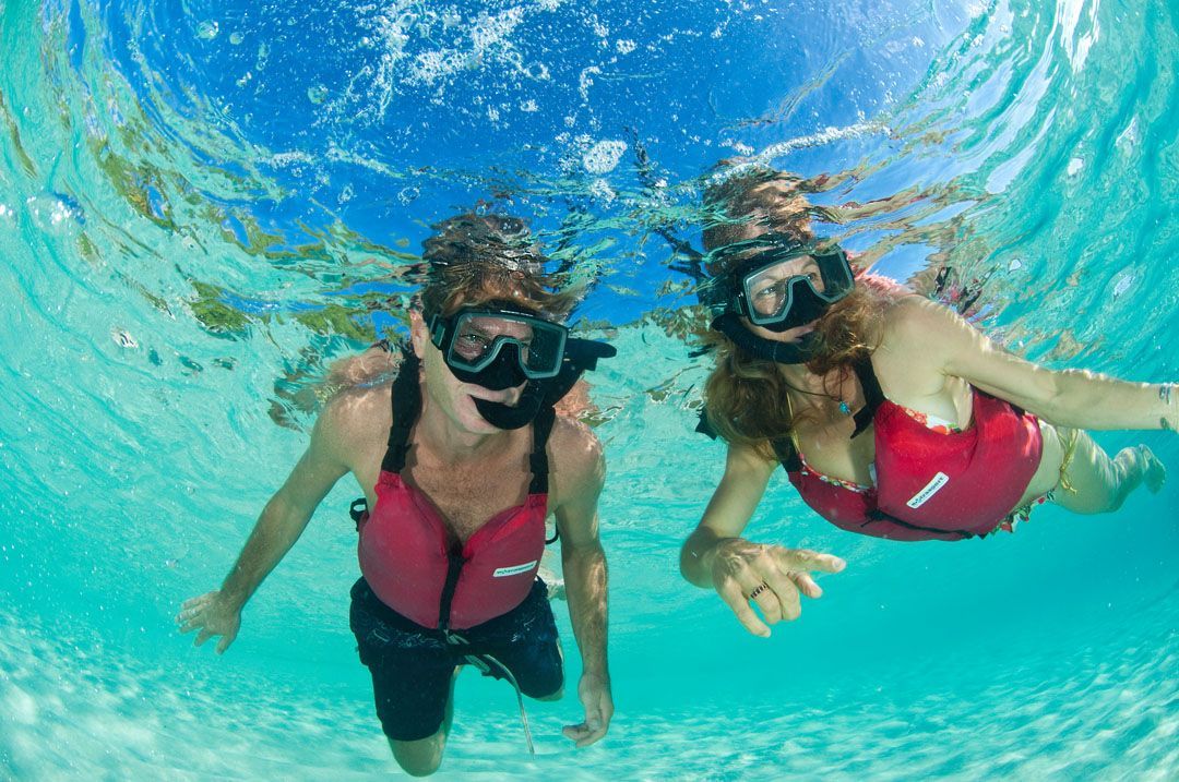 Two people snorkeling underwater, wearing masks and red life vests in turquoise water.