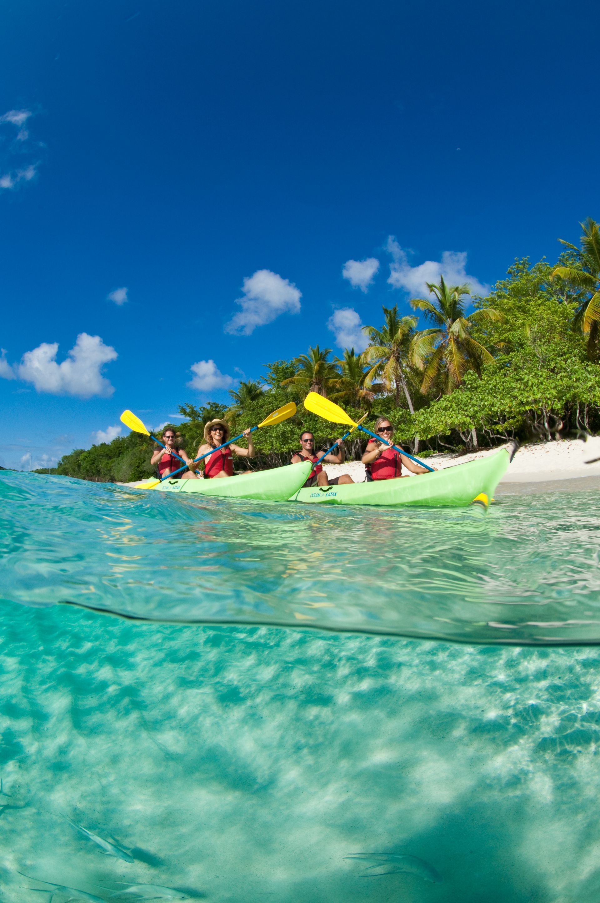 Kayakers on bright green kayaks paddle along a tropical beach with clear turquoise water and blue sky.
